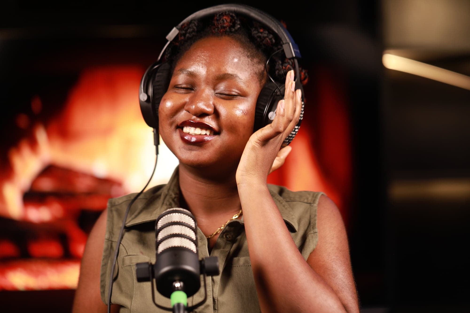 A smiling host in headphones at a RØDE PodMic, with the fireplace scene glowing behind her