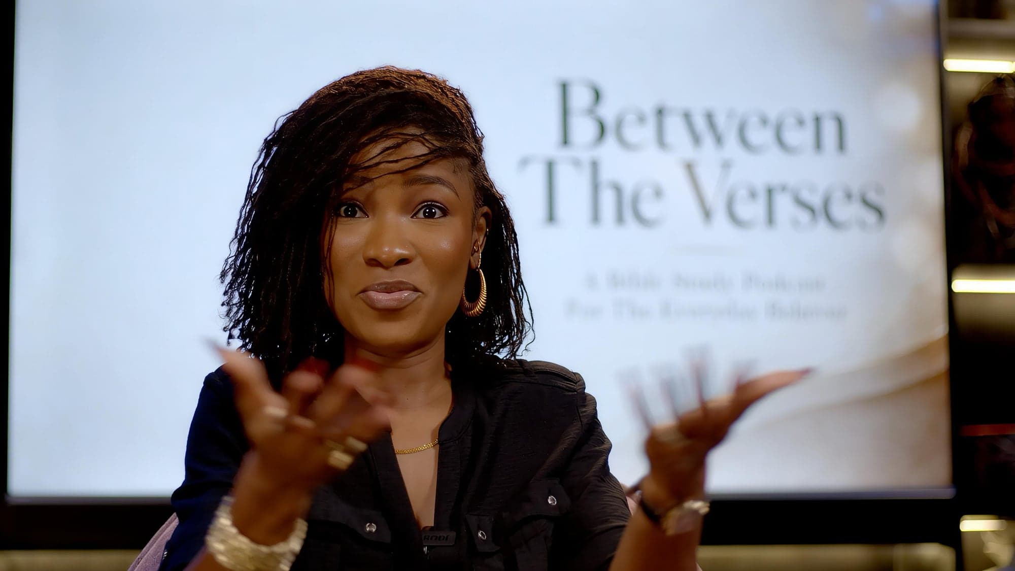 A host speaking on the Between The Verses set, with the show’s title lit on the 100-inch screen