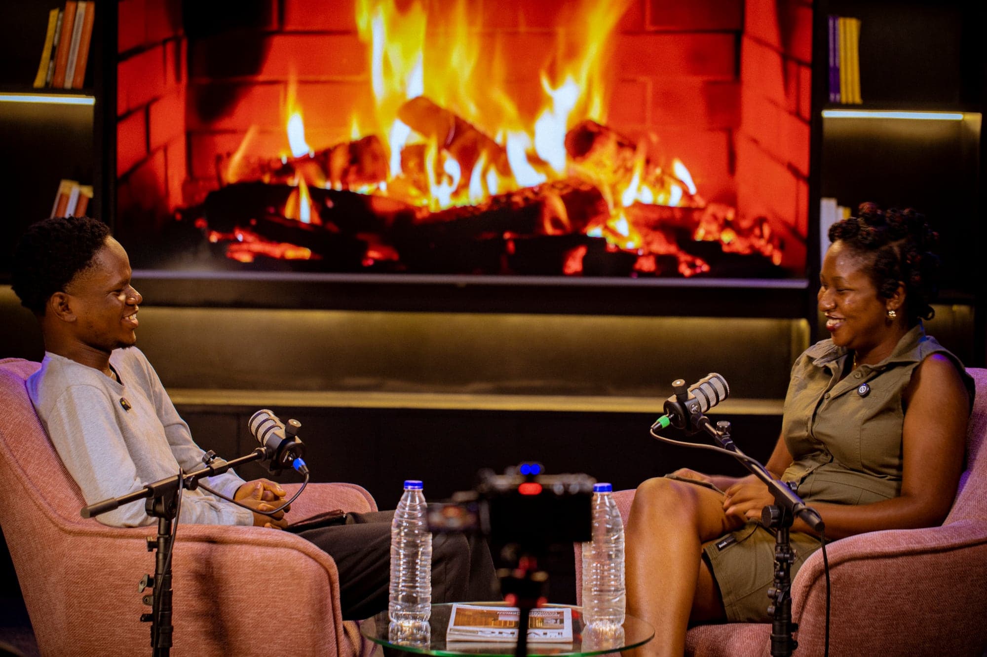 Two hosts laughing on the pink armchairs, with a fireplace scene lit on the 100-inch screen behind them