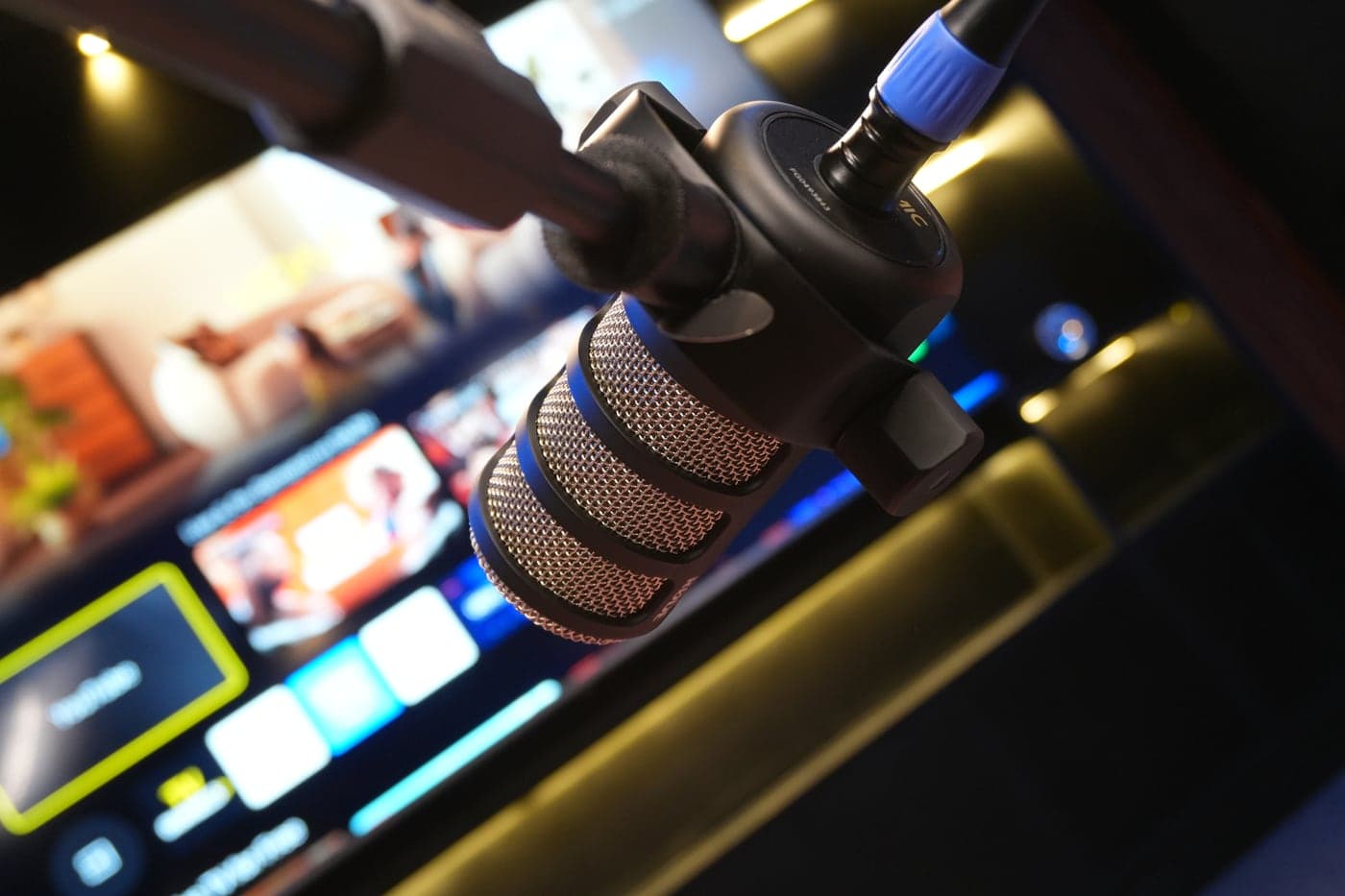 A RØDE PodMic on its boom arm, with the lit screen and shelves behind it